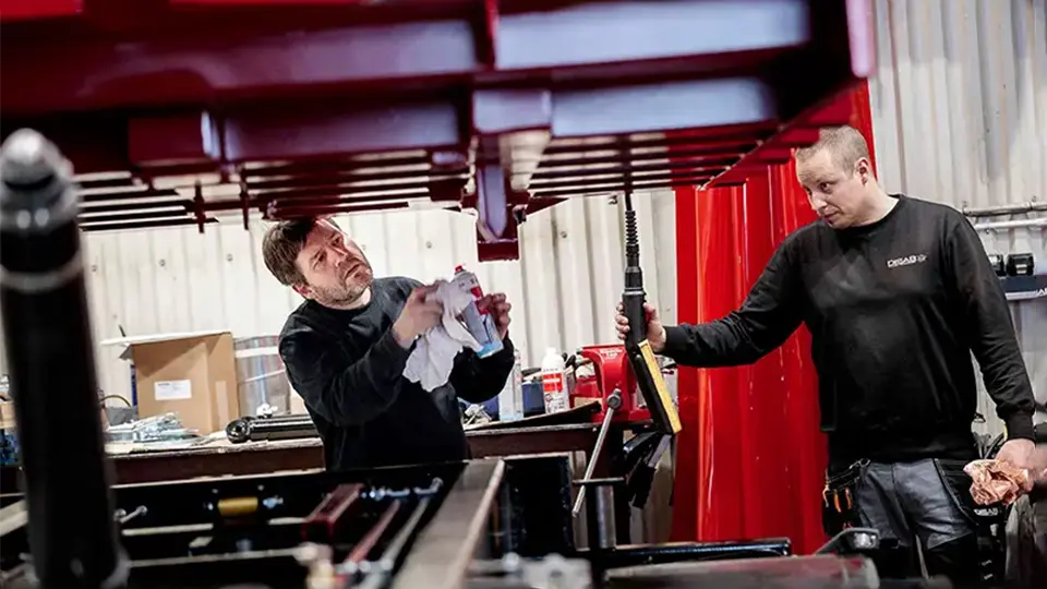 Två arbetare i svarta DISAB-uniformer utför underhåll på en röd industriell enhet, med hjälp av verktyg och rengöringsmedel i en välutrustad verkstad.
