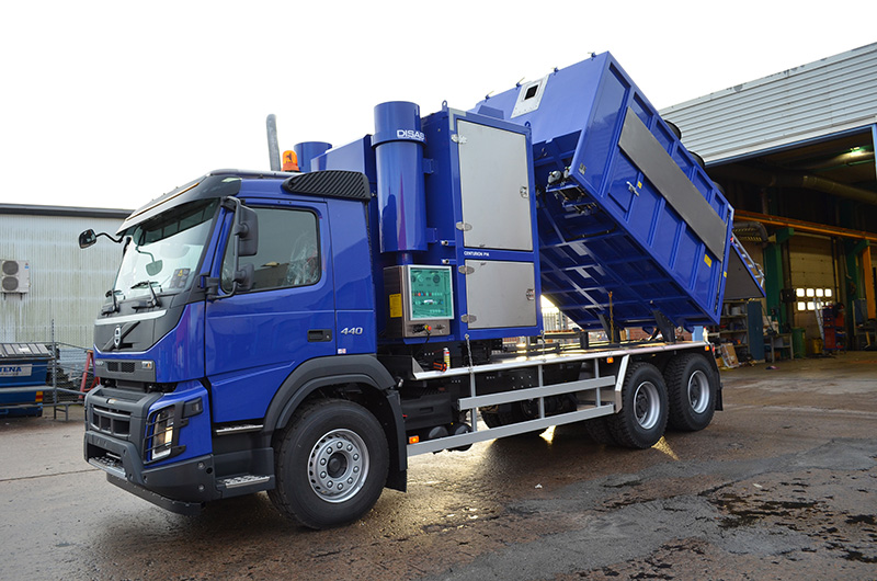 Blue DISAB Centurion™ P14 vacuum loader with its square tank lifted in a tipping position outside an industrial facility, demonstrating smooth hydraulic emptying for dry material handling.