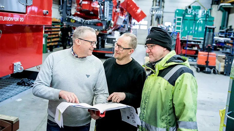 Tre män diskuterar en manual i en industriell verkstad, omgivna av DISAB vakuumutrustning, varav en bär en varseljacka.
