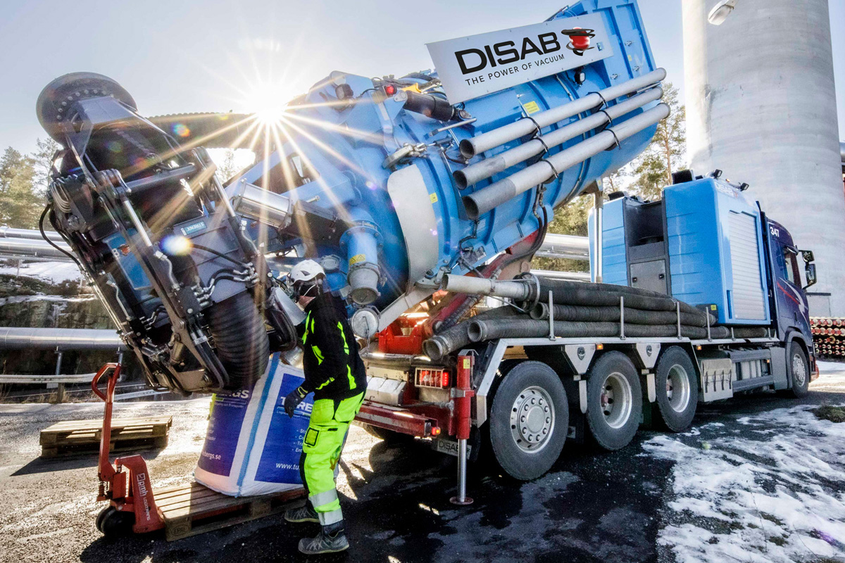 DISAB blue vacuum loader truck tips its tank, emptying collected material into a pallet bag while a worker supervises at an industrial site.