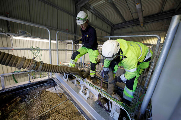 Two operators in safety gear manage an industrial vacuum hose connected to a DISAB unit, extracting dry material at a worksite.