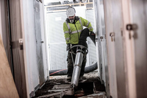 Operatör i skyddsutrustning använder en stor sugslang från en DISAB-enhet inuti en byggnad, vilket betonar säkerhet, ergonomi och materialhantering i trånga industriella utrymmen.
